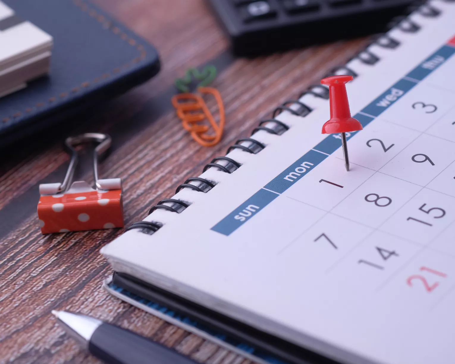 A picture of a desk calendar on a wood desk with a red push-pin in the first of the month. A binder clip, part of a pen, and part of a keyboard can be seen on the desk surrounding the desk calendar.