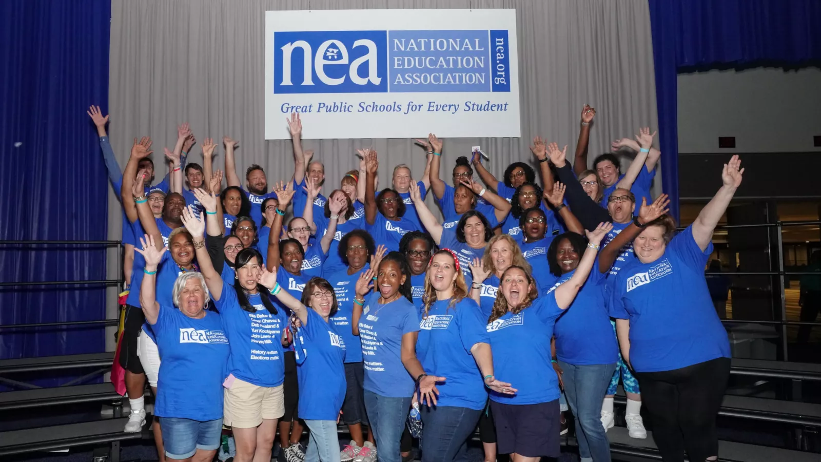 DSEA members elected to serve in the 2023 NEA RA delegation pose for a fun picture together with their hands in the air underneath an NEA banner.