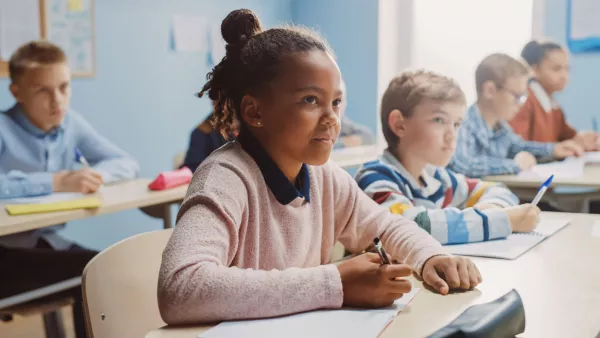 a black girl in a pink sweater sits at a desk holding a pencil, surrounded by other students