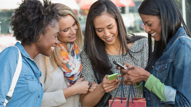 Four women shopping together look at two phones with excitement.
